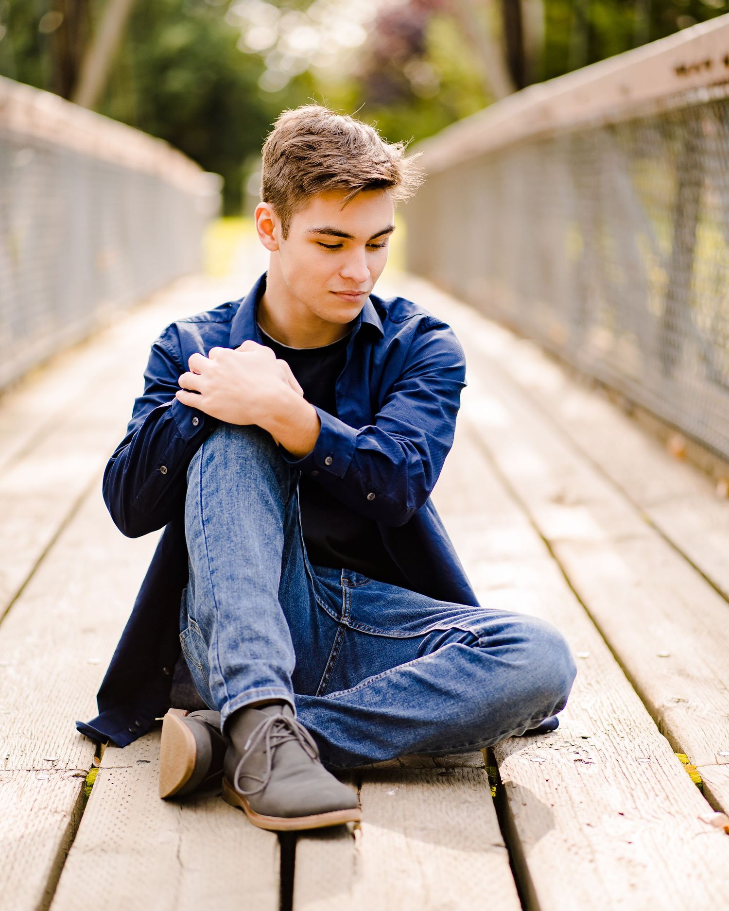 A high school boy in casual wear sits on a wooden bridge in an outdoor setting with natural lighting for his senior photos in Tacoma.