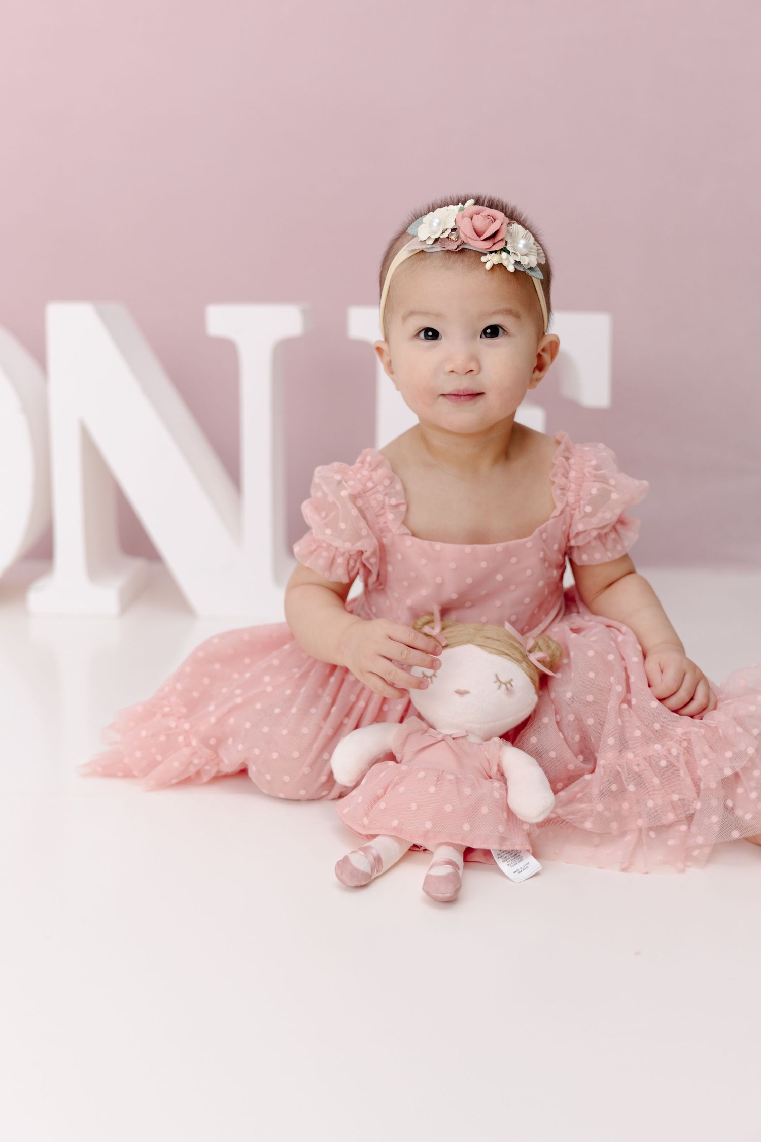 Sweet baby in pink lace dress with floral headband sits on white backdrop in professional photo session.