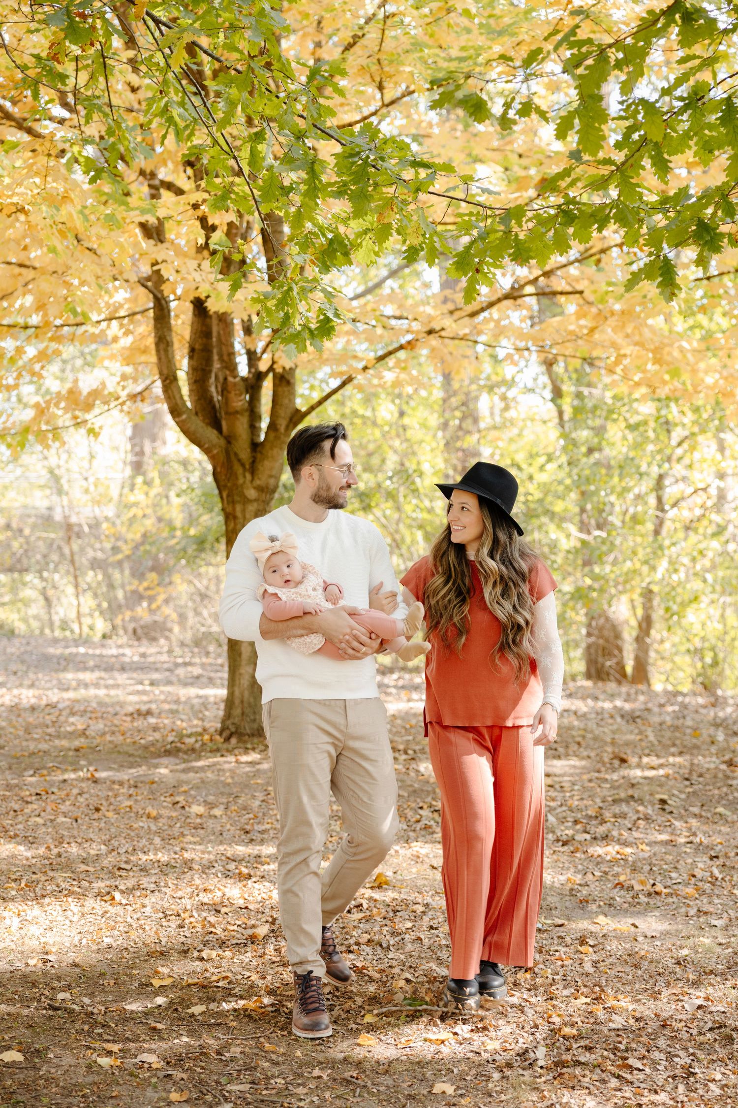 A couple walks together with their baby through a scenic autumn forest with golden leaves and dappled sunlight.