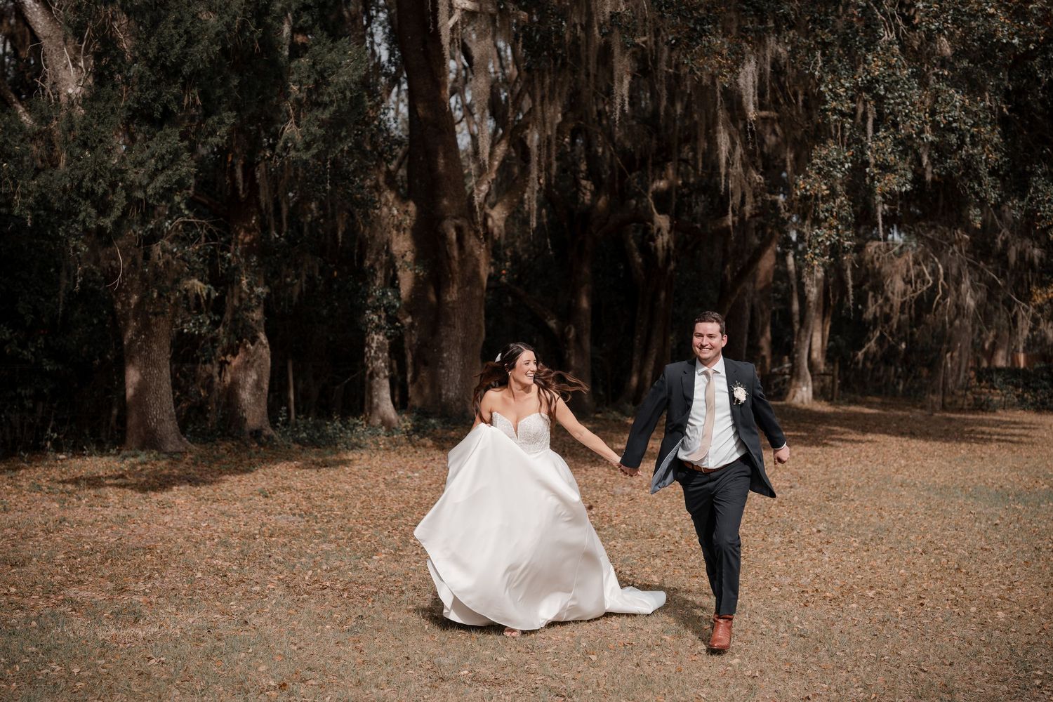A couple runs joyfully through a path lined with Spanish moss-draped oak trees in their wedding attire.