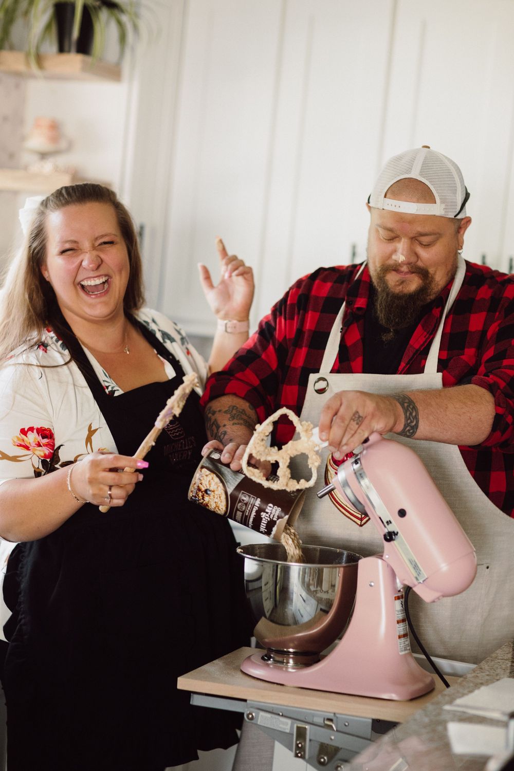Two people having fun while baking with a pink KitchenAid mixer in a bright white kitchen.