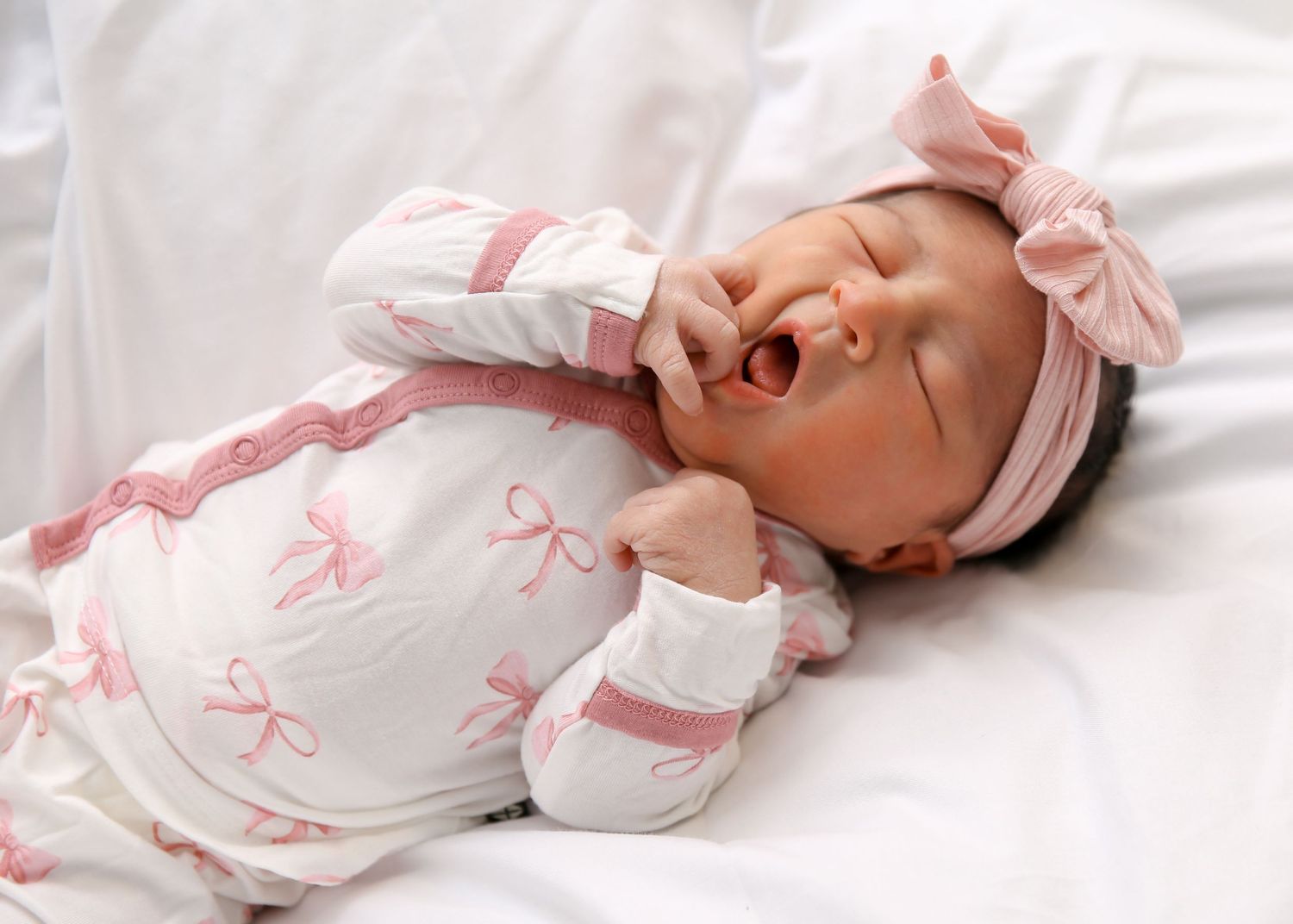 Baby wearing pink and white outfit lying on white bedding smiling and playing with hands and feet.