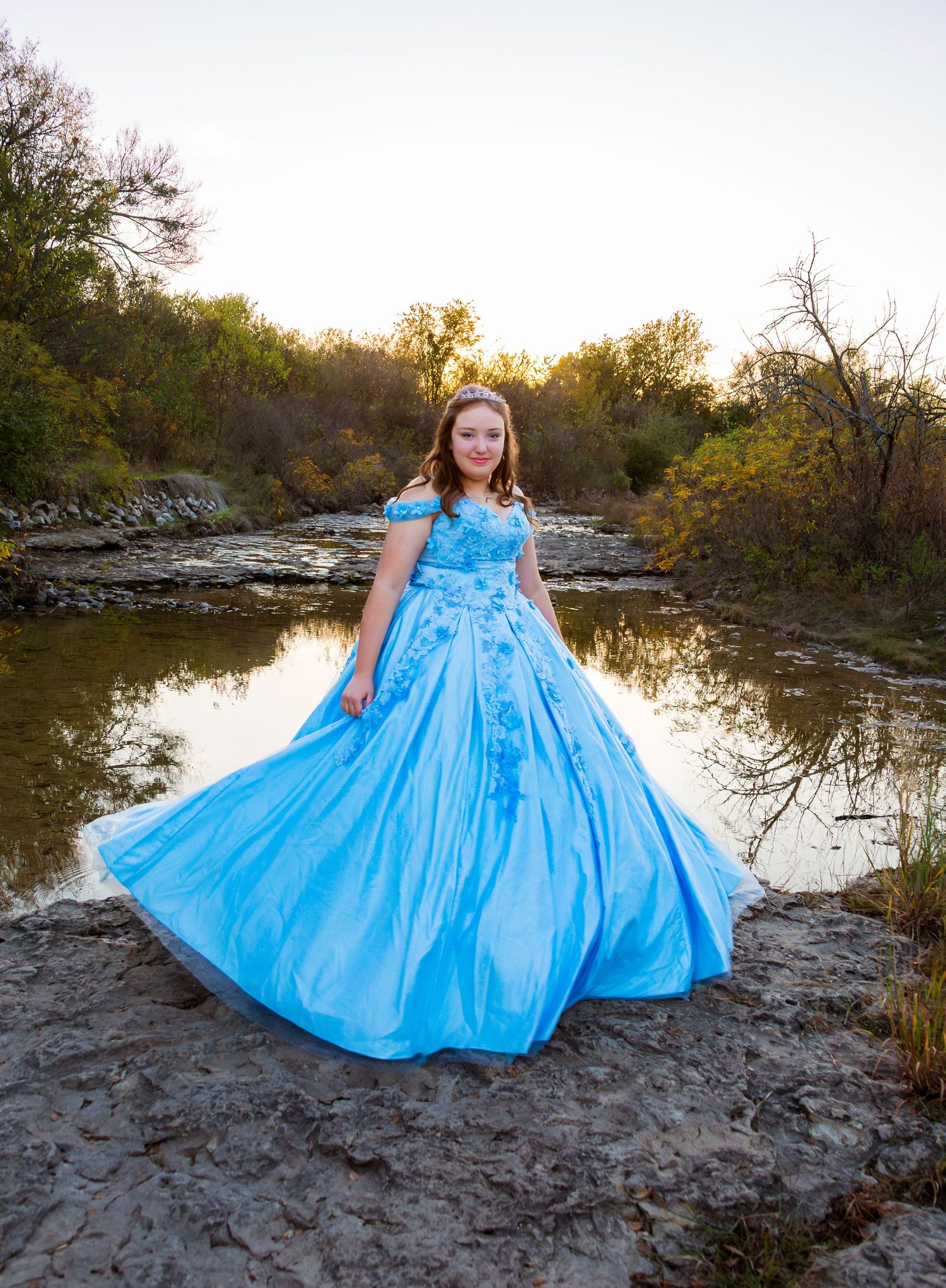 A person in a flowing blue ball gown poses against a scenic desert landscape at sunset.
