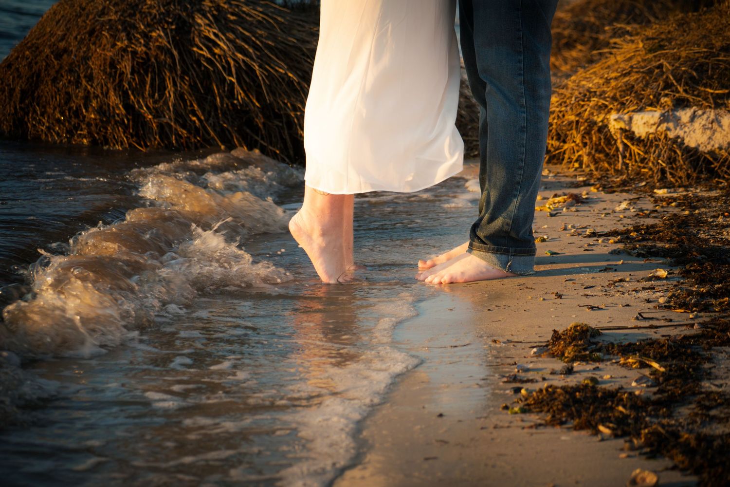 Bare feet stand together in shallow ocean waves on a sandy beach at sunset.