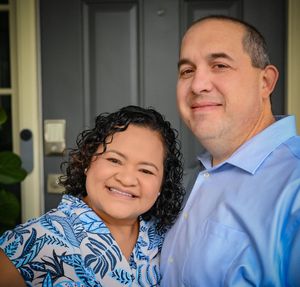 A couple in coordinating blue outfits smile warmly together in an outdoor portrait photo.