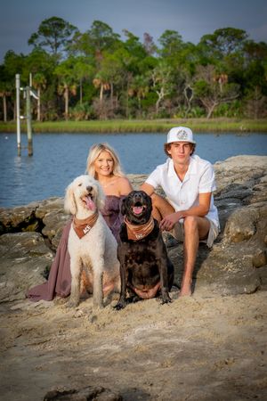 Three dogs and their companion pose together on rocky terrain near a waterfront with trees in the background.