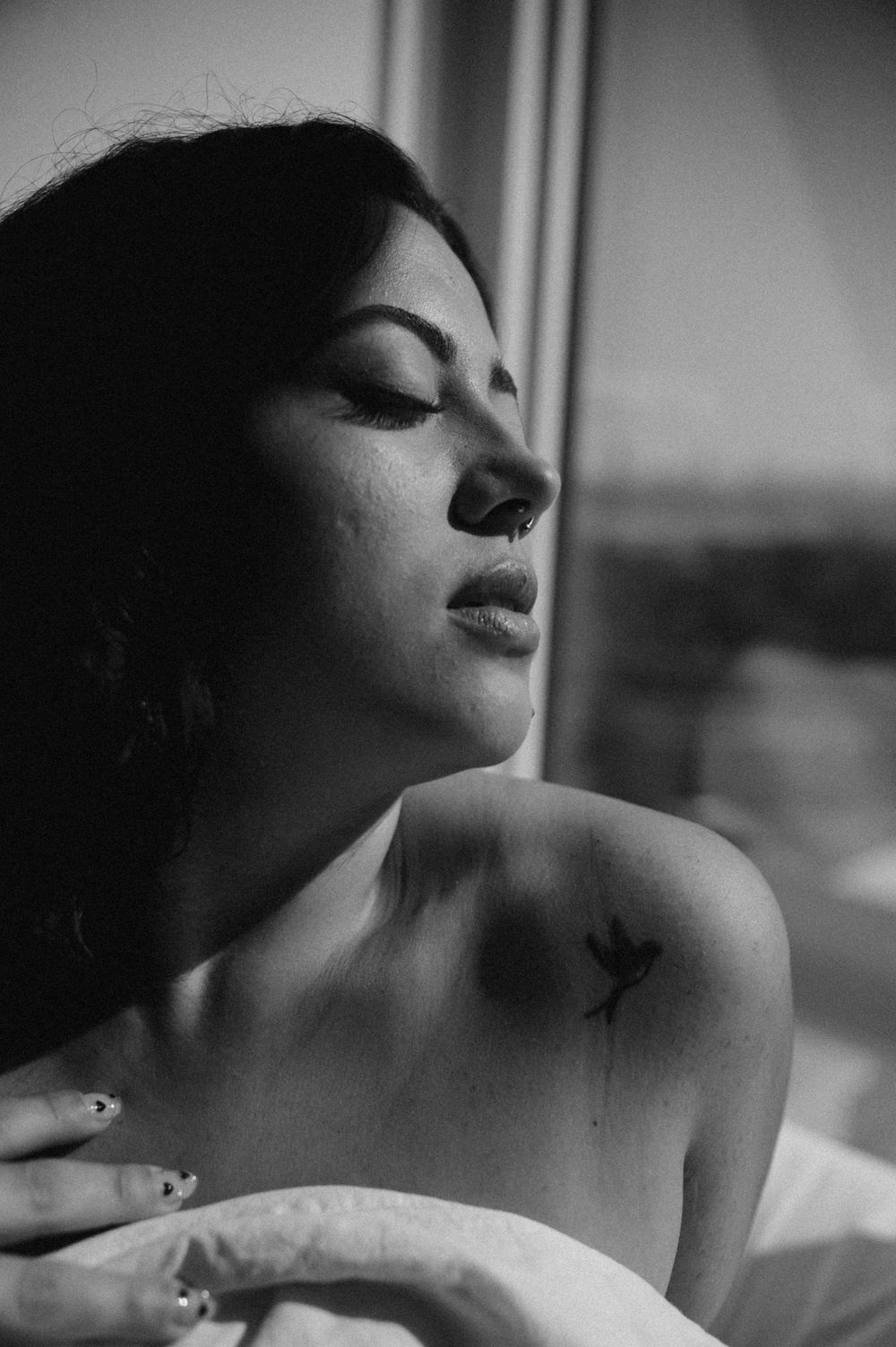 Black and white portrait showing a small bird tattoo on shoulder near window.