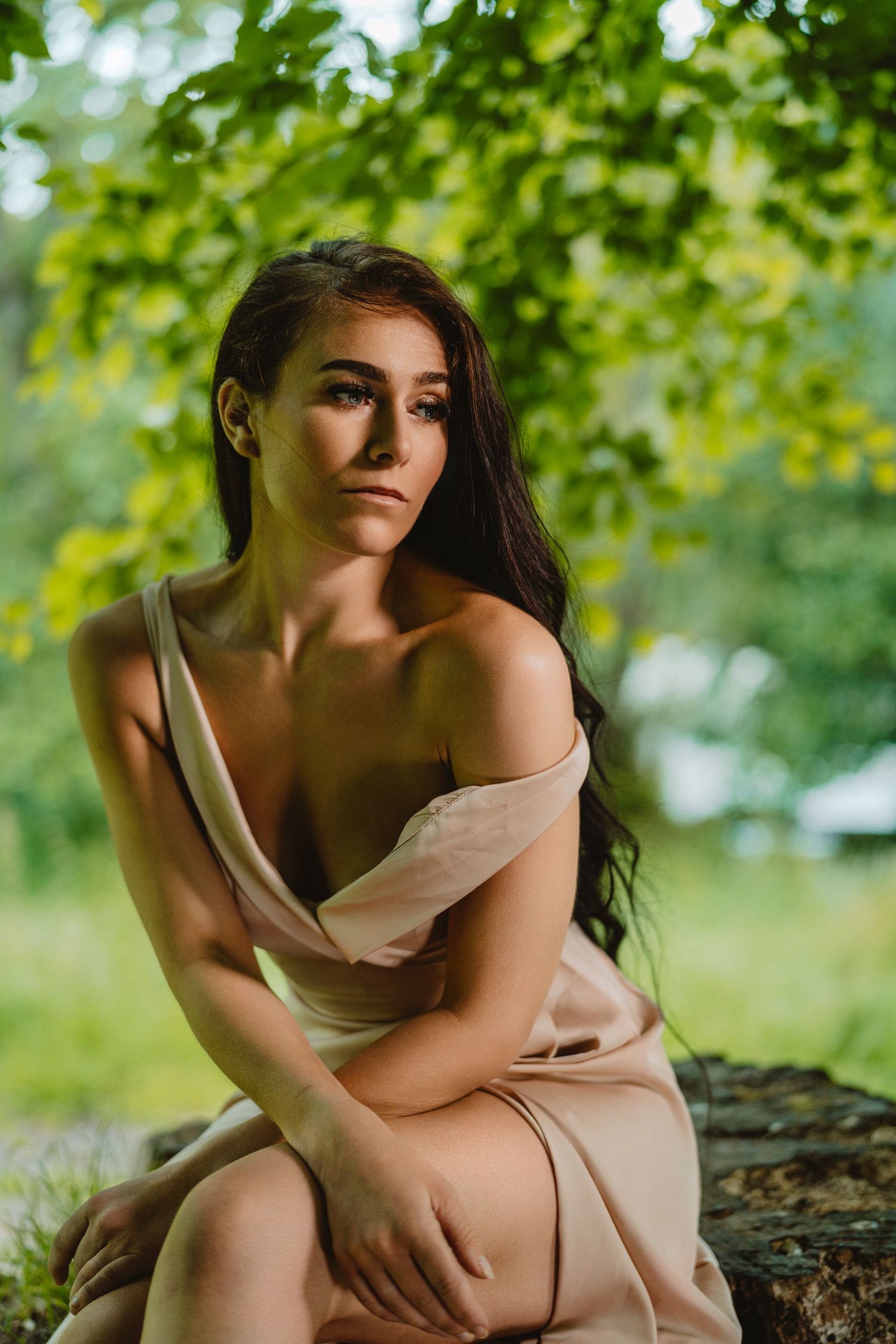 A person in a beige dress poses outdoors under green foliage in soft natural lighting.