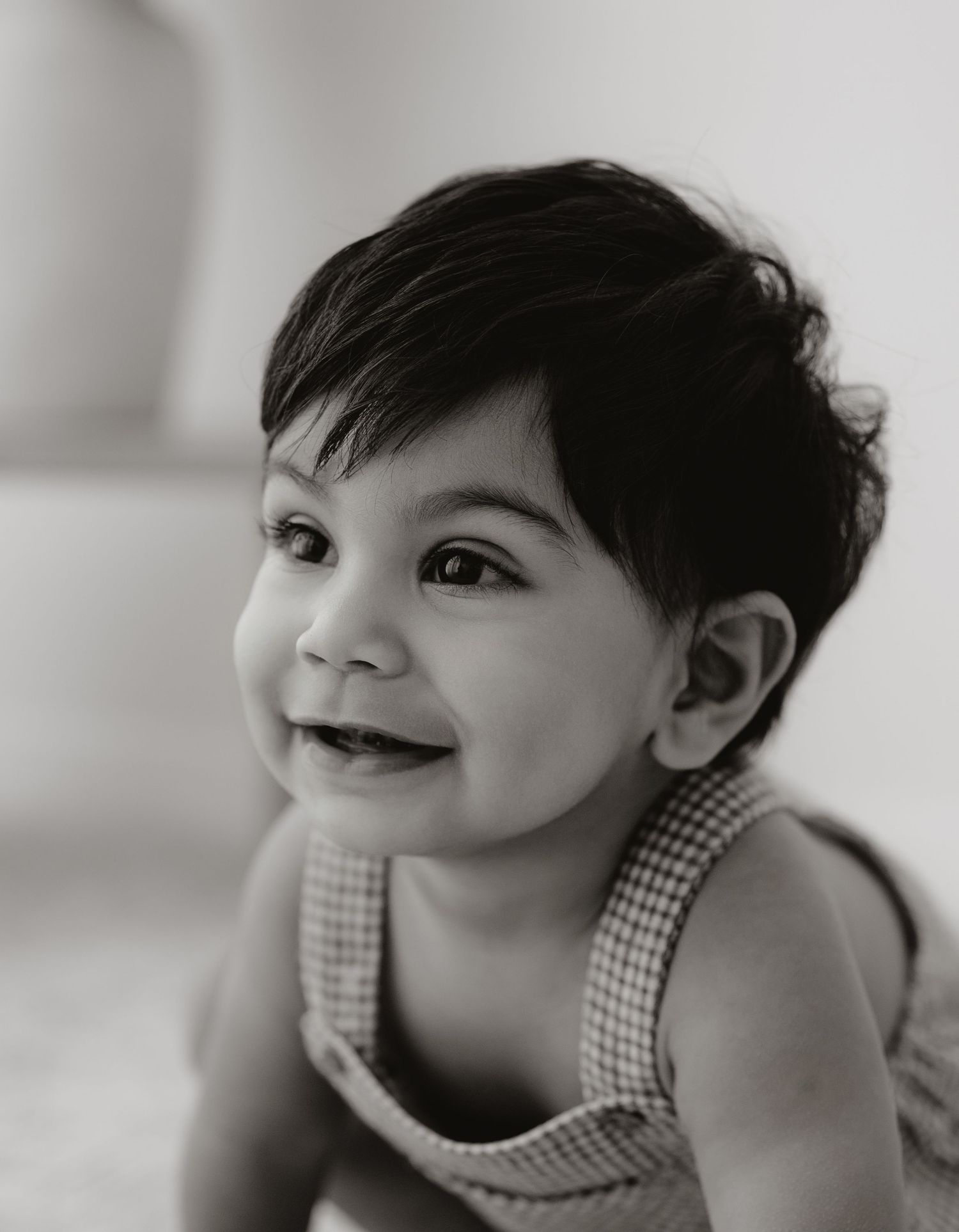 Baby in white dress plays peek-a-boo under cream blanket on textured bedding in warm lighting.