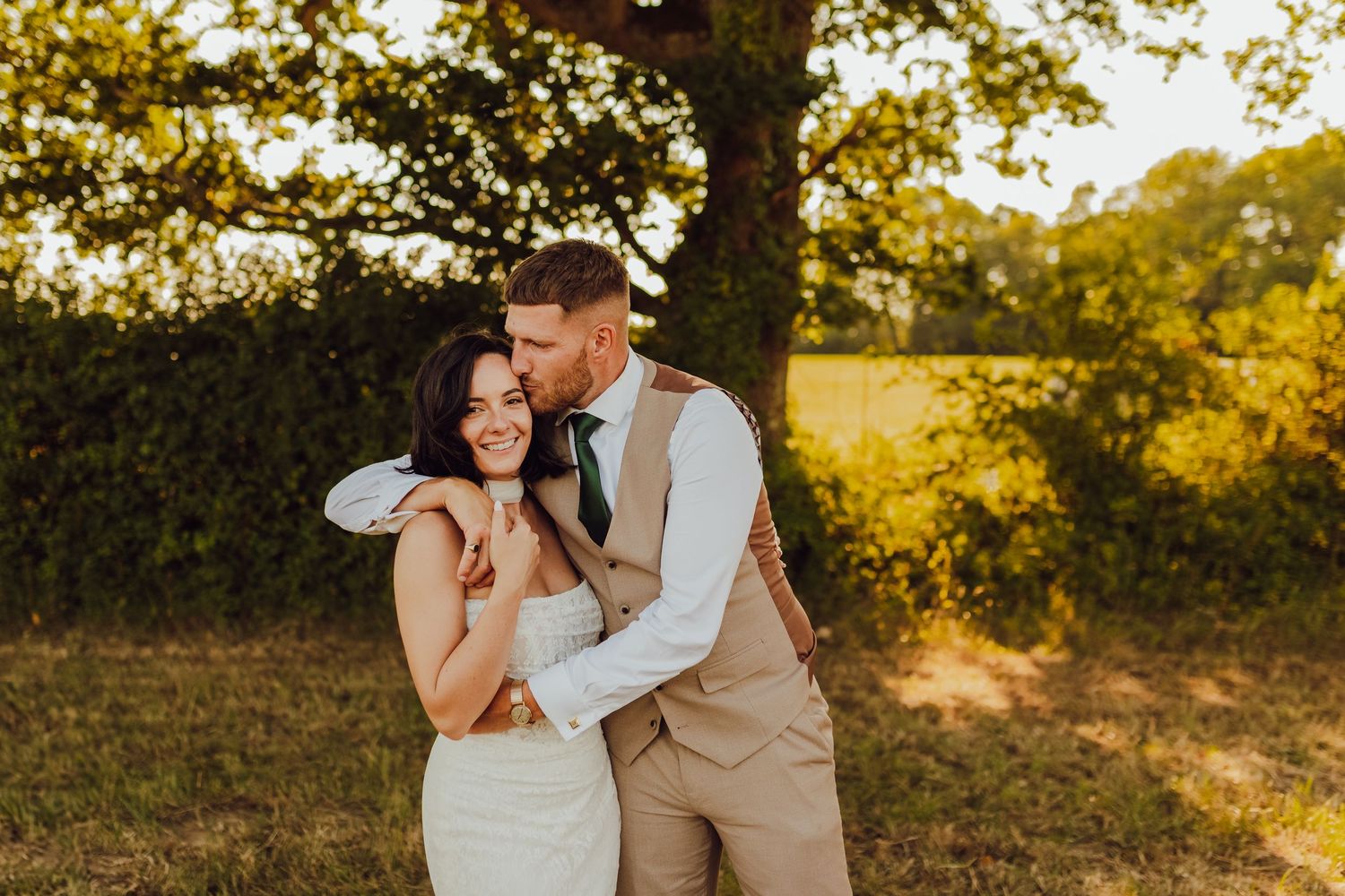 The wedding couple posing together in a field during golden hour, embracing and smiling.