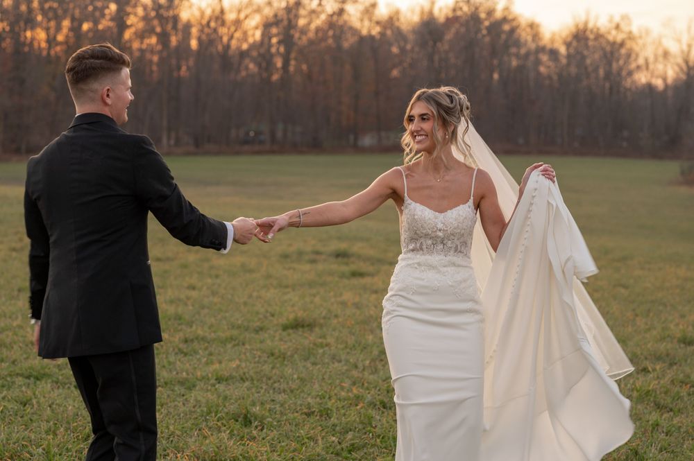 A bride in a black dress and veil poses playfully in a grassy field at sunset.
