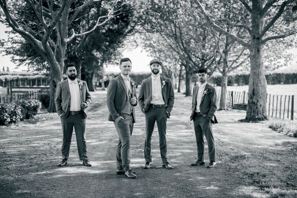Four men in formal attire pose casually under trees in a black and white photograph. Hamilton photographer