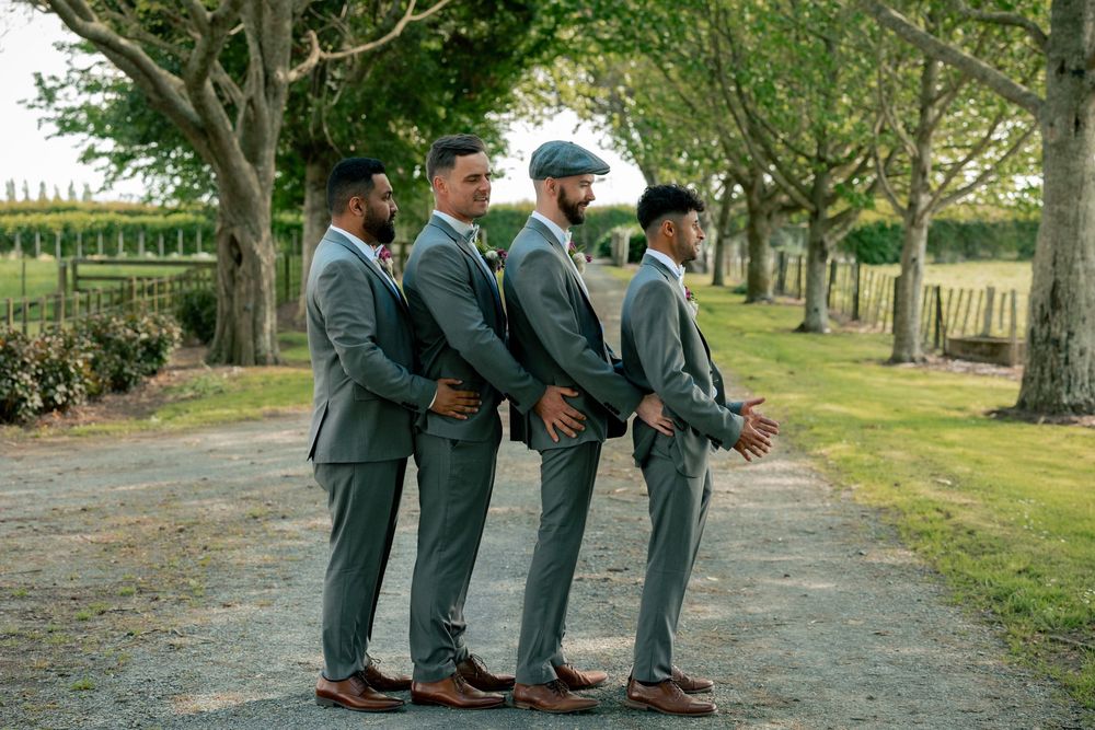 Groomsmen in matching dark suits posing together on a tree-lined path.