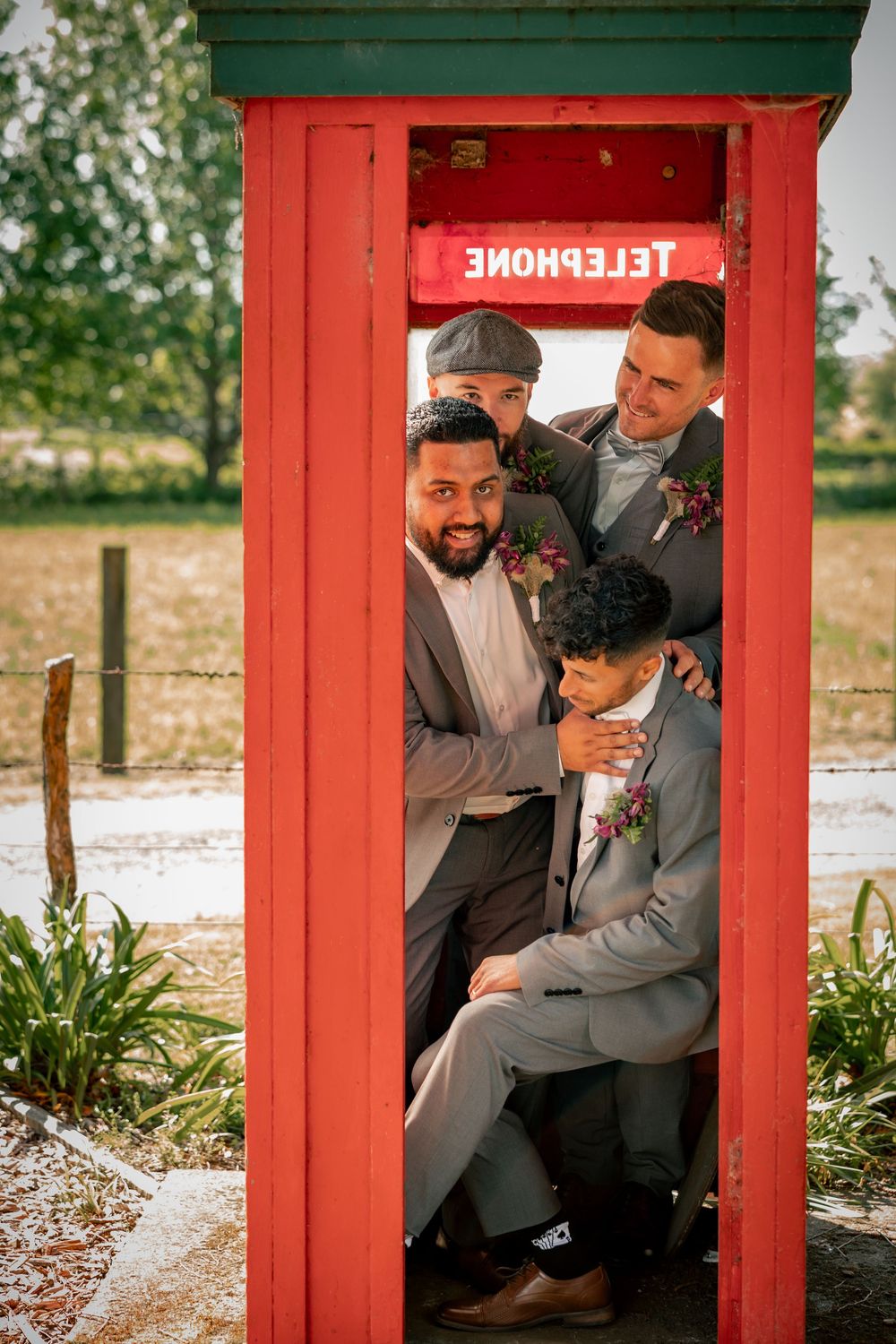 A group of people in formal attire squeeze into a bright red British telephone booth for a fun wedding photo.