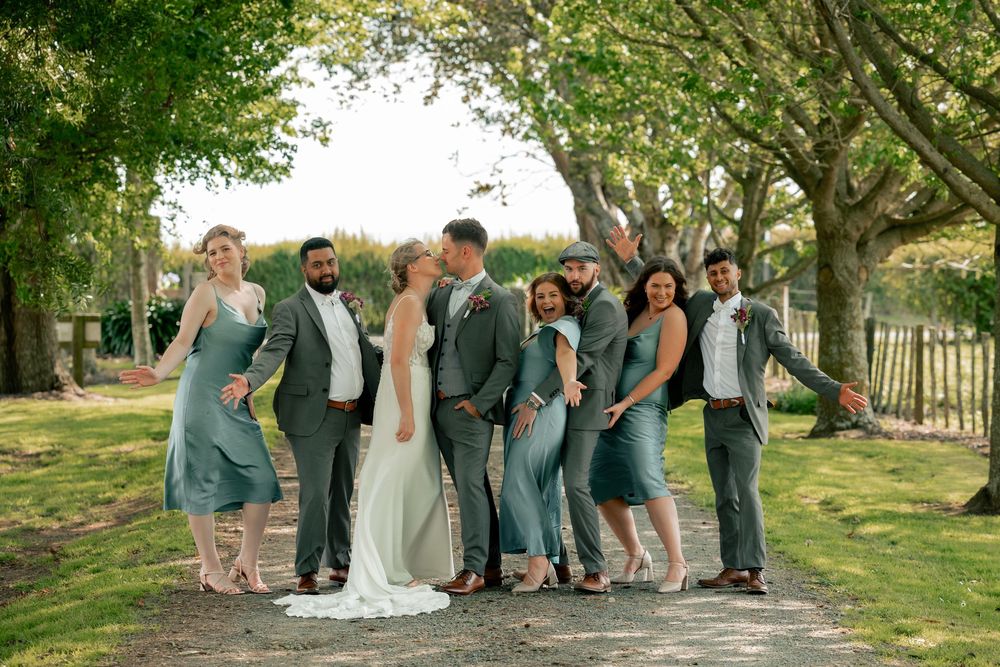 Wedding party members gathered outdoors under green trees for group photos.