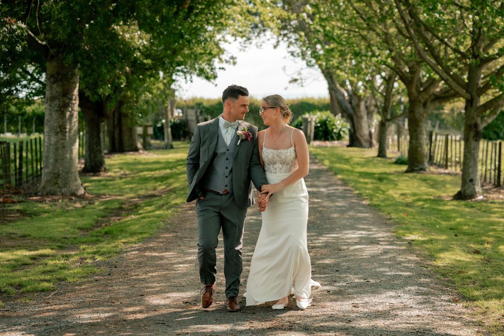 A couple walks along a tree-lined path in a garden setting during a wedding photoshoot.