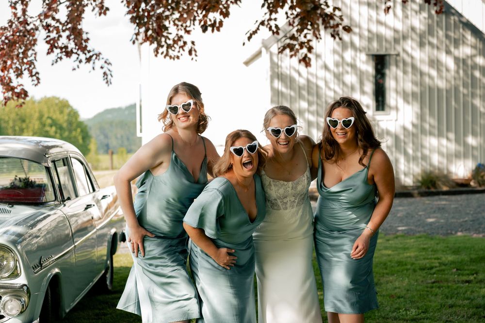 A group in matching teal bridesmaid dresses and sunglasses pose together by a vintage car on a sunny day.