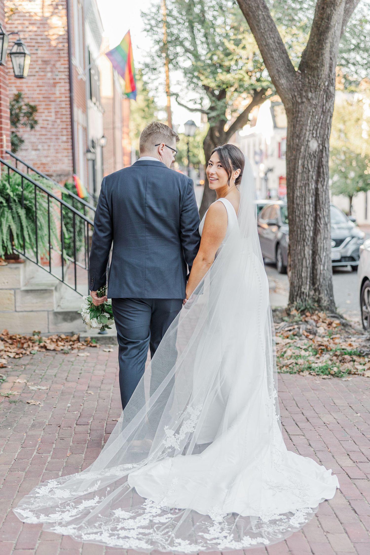 A wedding couple walks down a brick pathway with trees and buildings in the background, her long lace veil trailing behind.