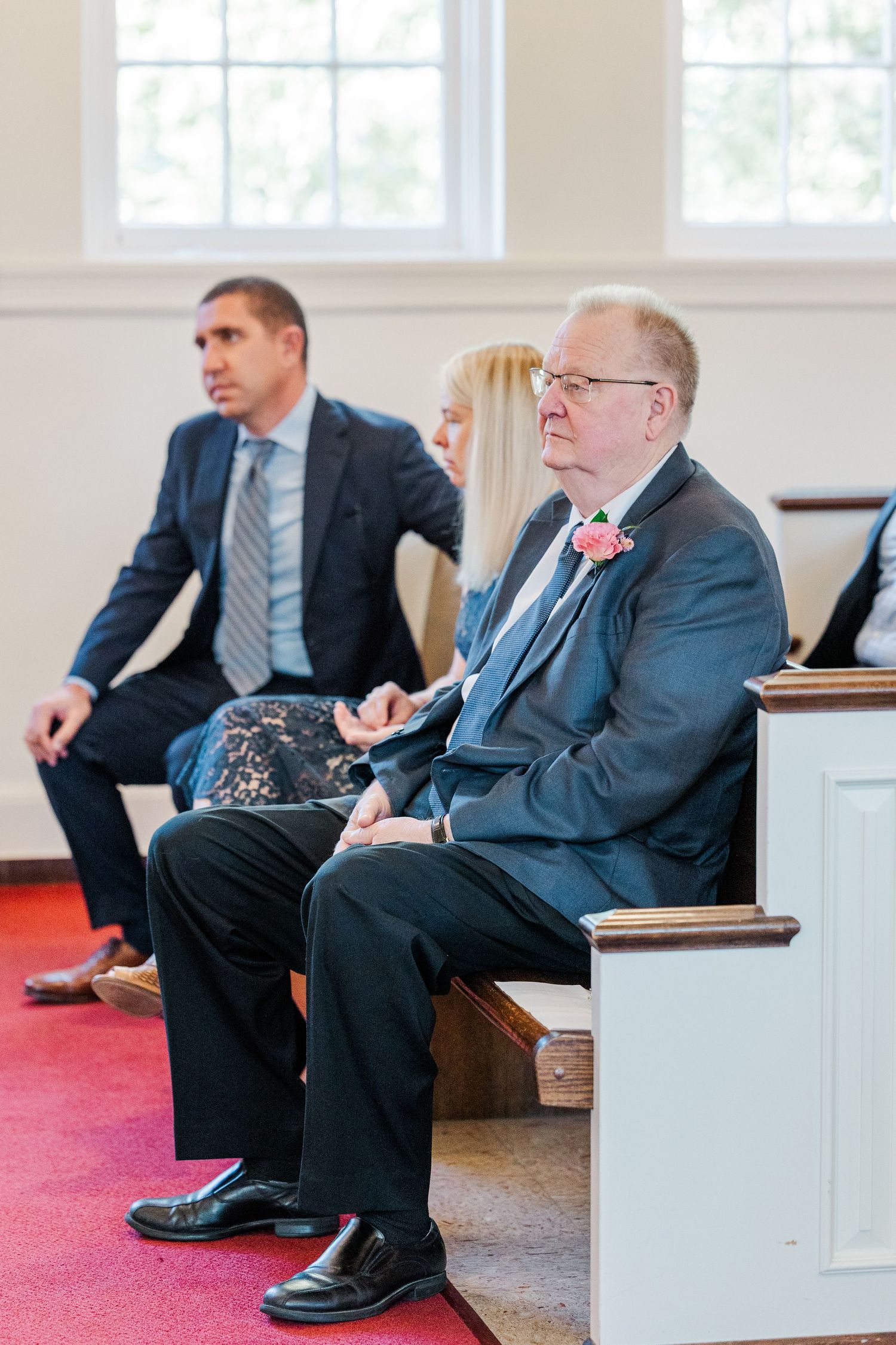 Several people in formal attire sit on church pews with red carpeting during a ceremony.