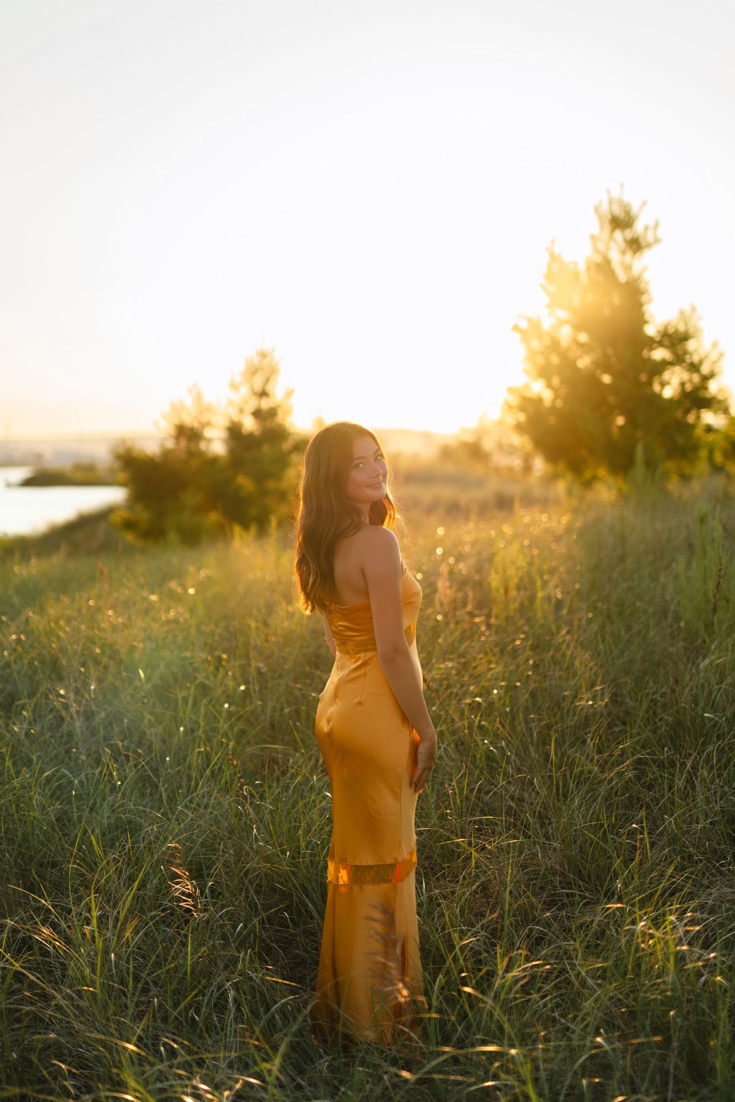 A person in a flowing yellow dress stands in a sunlit meadow during golden hour as tall grass sways in the breeze.