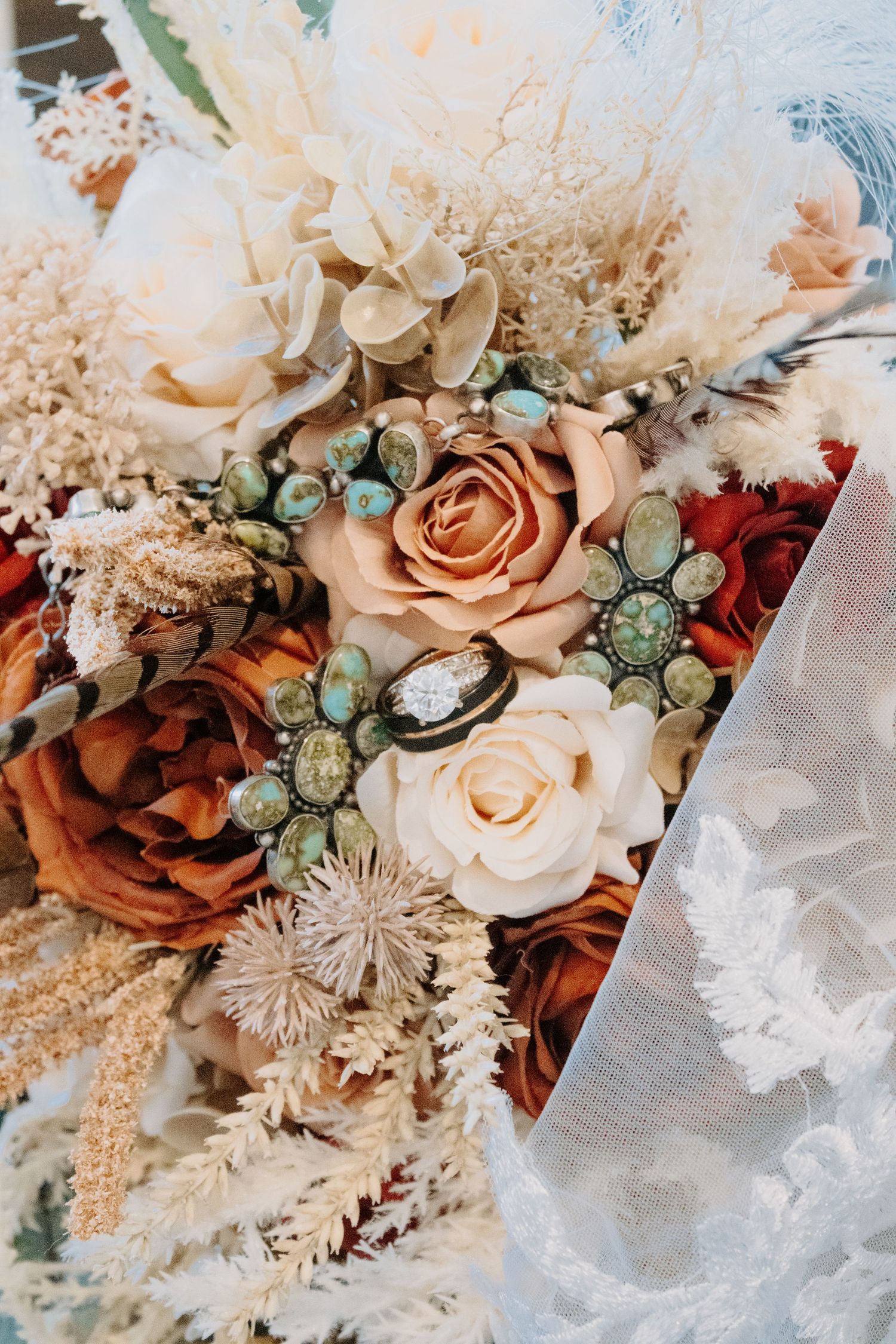Boho wedding bouquet with rust and cream roses, dried pampas grass, and turquoise jewel accents beside a lace veil.