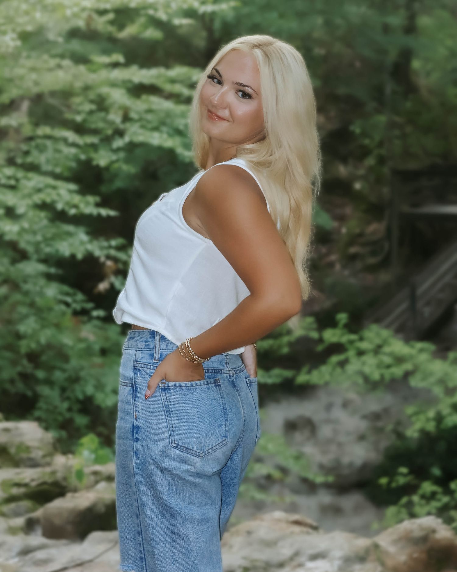 A person posing casually against a natural backdrop of green foliage.