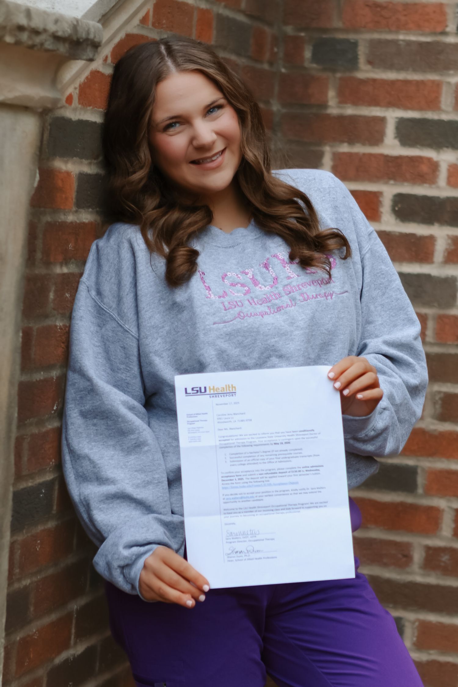 Person wearing gray sweatshirt shows official document while seated by brick wall.