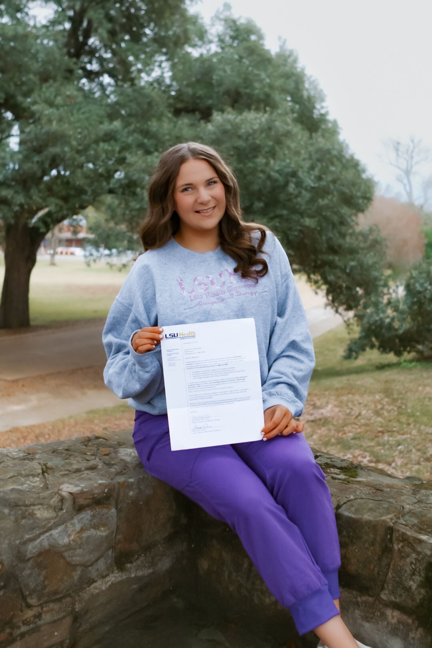 Person in gray top and purple pants displays document while sitting outdoors.