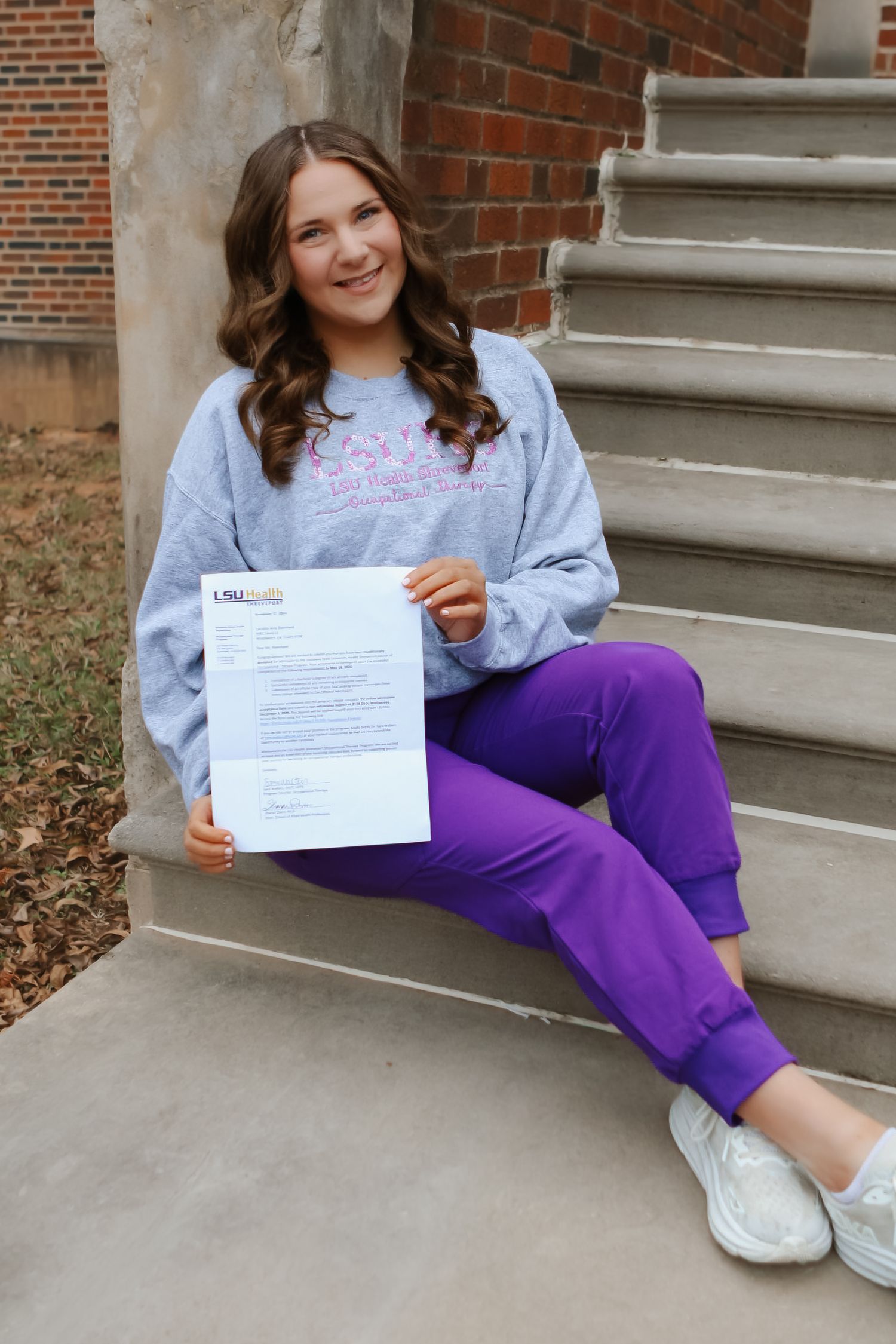 Person in gray sweatshirt and purple pants sits on concrete steps holding document.