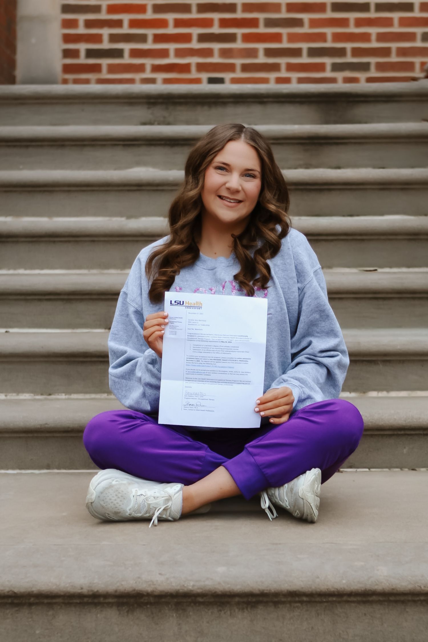 Person in casual attire displays LSU document while sitting on brick building steps.