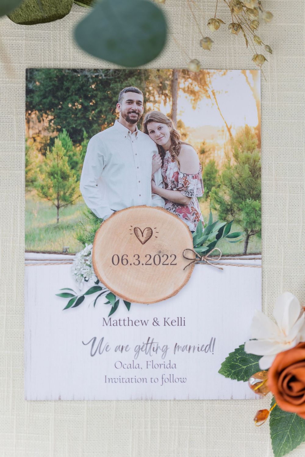 Save the date wedding invitation featuring a wooden disc with date and greenery details on a light background.