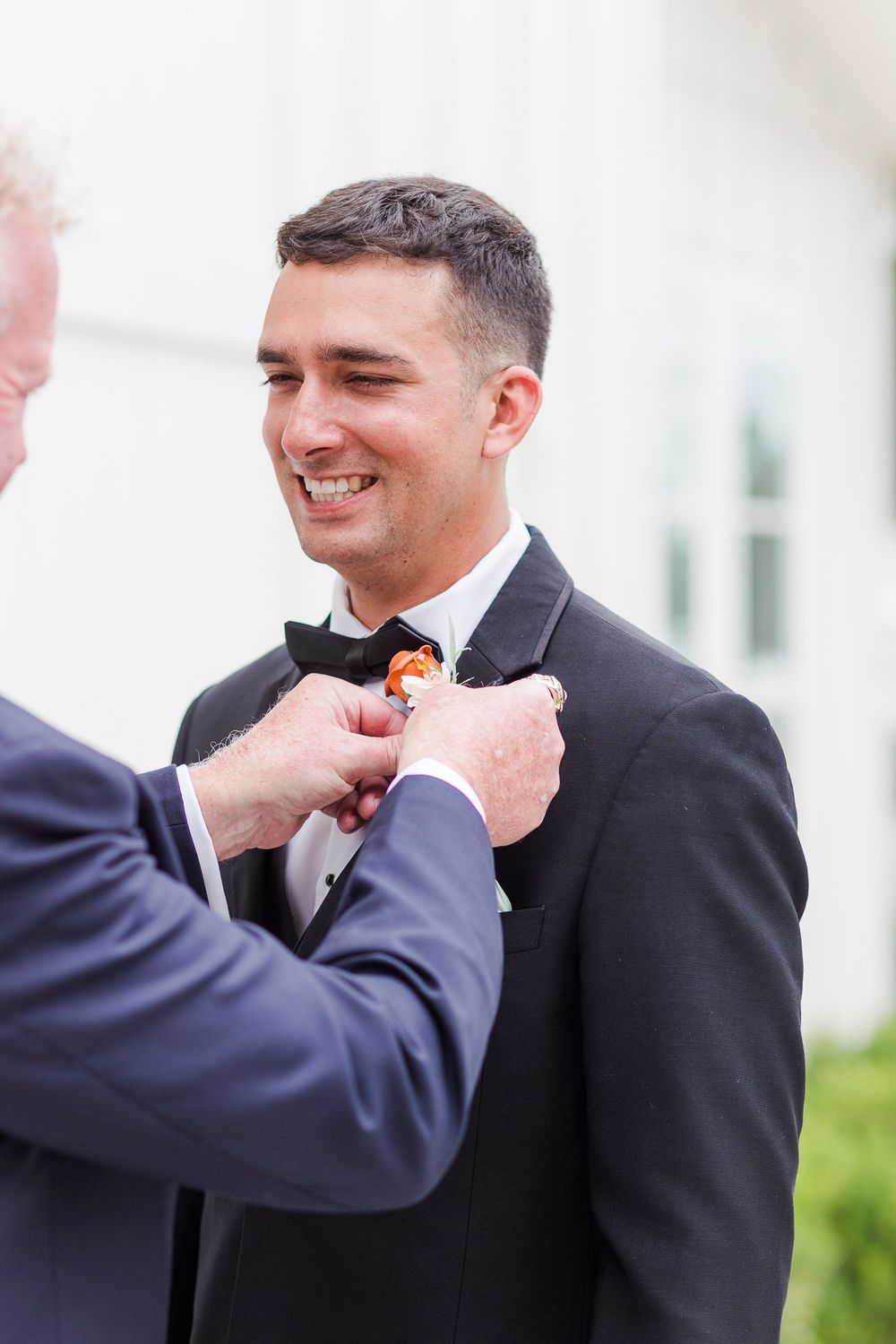 A groom smiles while someone adjusts his boutonniere during wedding preparations.