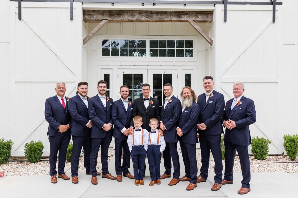 Wedding party in navy suits and two ring bearers pose in front of white barn.