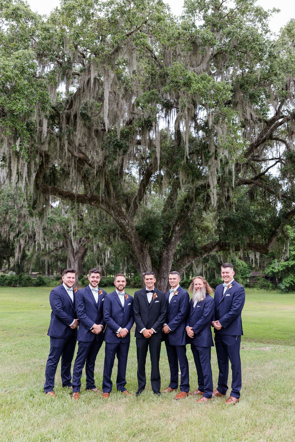 Groomsmen in navy suits lined up under moss-draped oak trees for outdoor wedding photo.