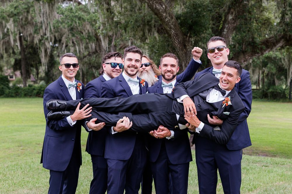 Groomsmen in navy suits pose playfully outdoors under Spanish moss draped trees.