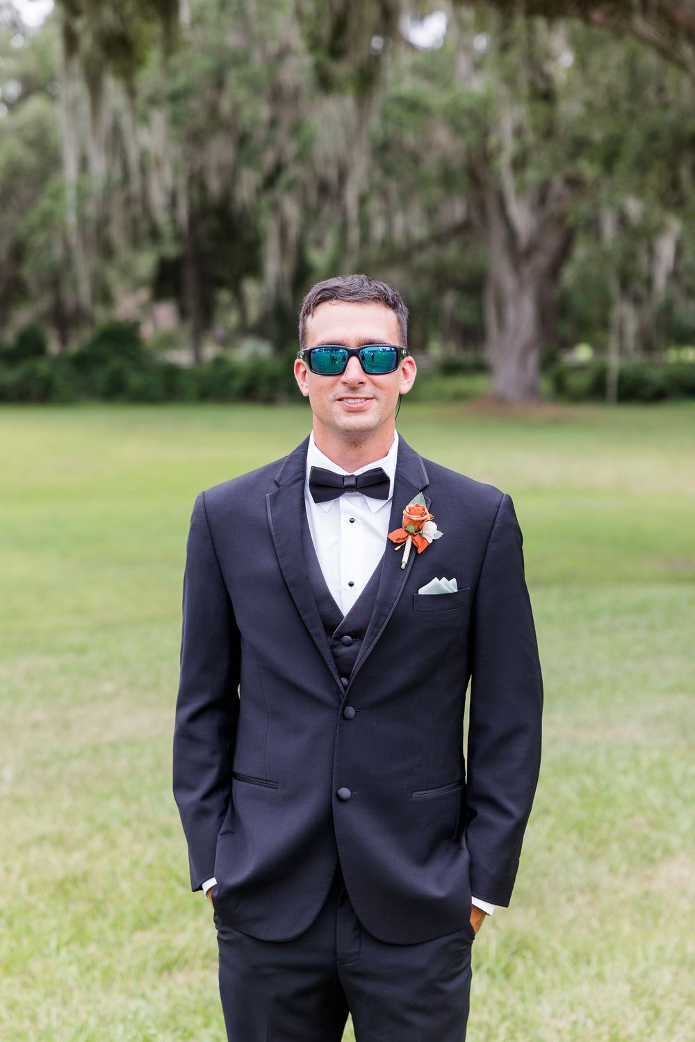 Groomsmen in black tuxedos and bowties stand on grass with trees in the background.