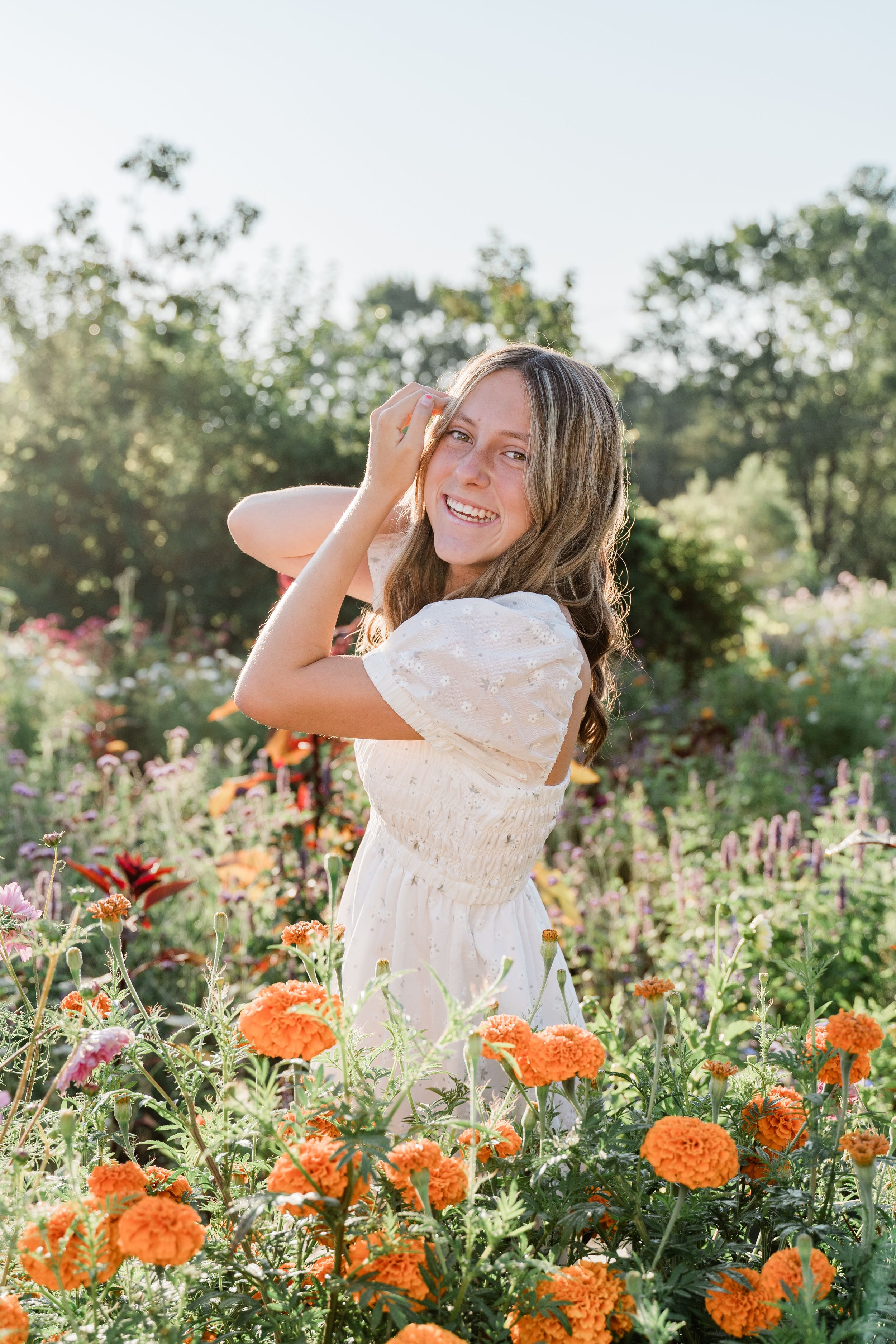 A person in a white dress poses joyfully among vibrant orange marigolds in a sunny garden setting.