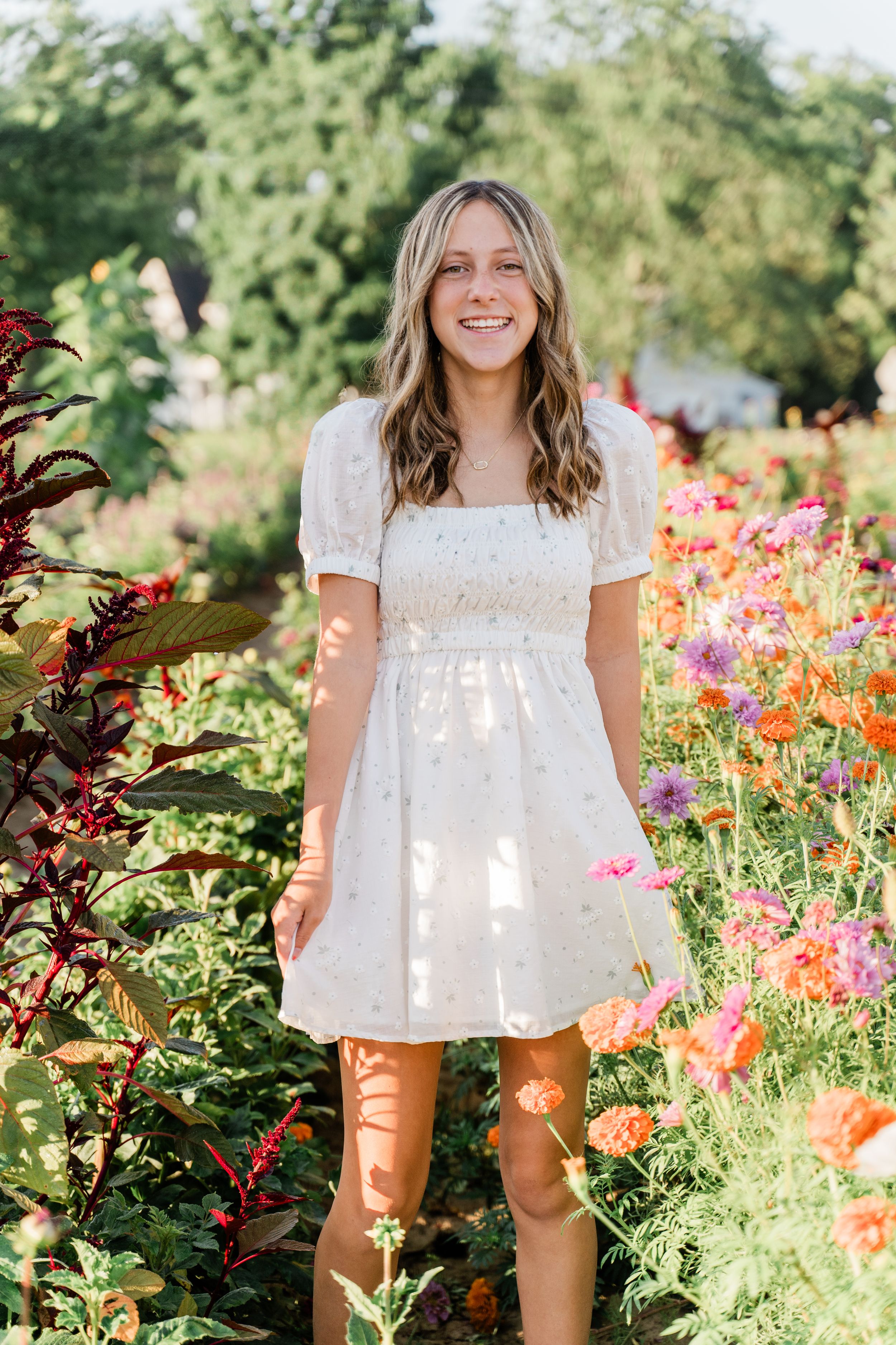 A white eyelet lace dress is captured in a sunny garden filled with vibrant orange and pink flowers.