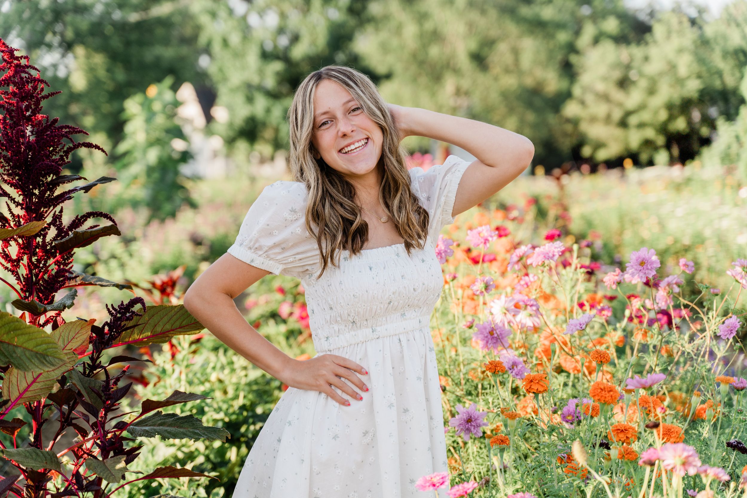 A person wearing a white lace dress stands cheerfully in a colorful summer garden with mixed flowers.