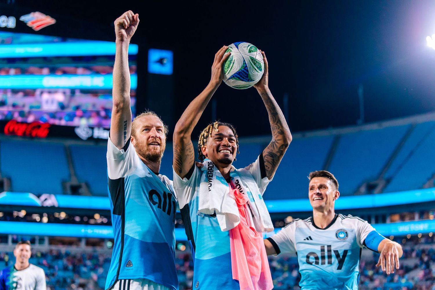 Two MLS soccer players celebrate a victory by holding up a match ball on the stadium field.