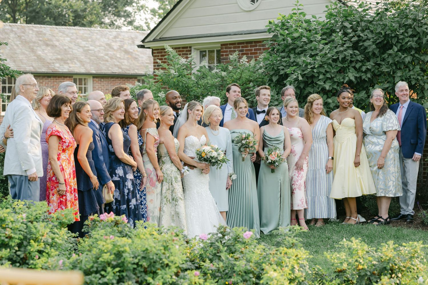 Wedding party members in pastel dresses and formal attire stand together in a garden setting for group photos. - Elawa Farm
