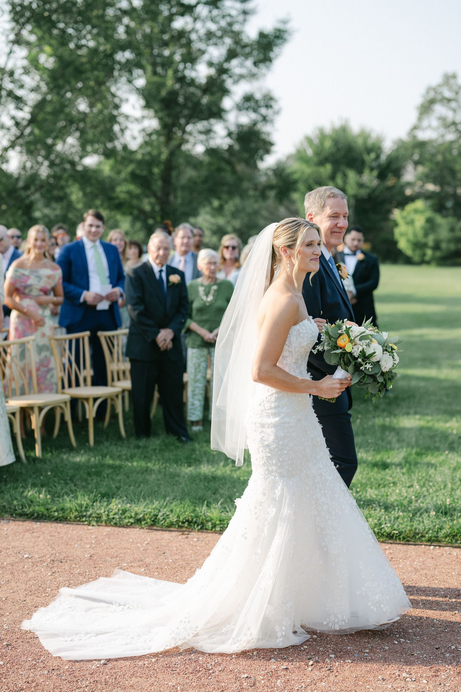 Bride in flowing white dress dances with partner on an outdoor path during golden hour with guests watching. - Elawa Farm
