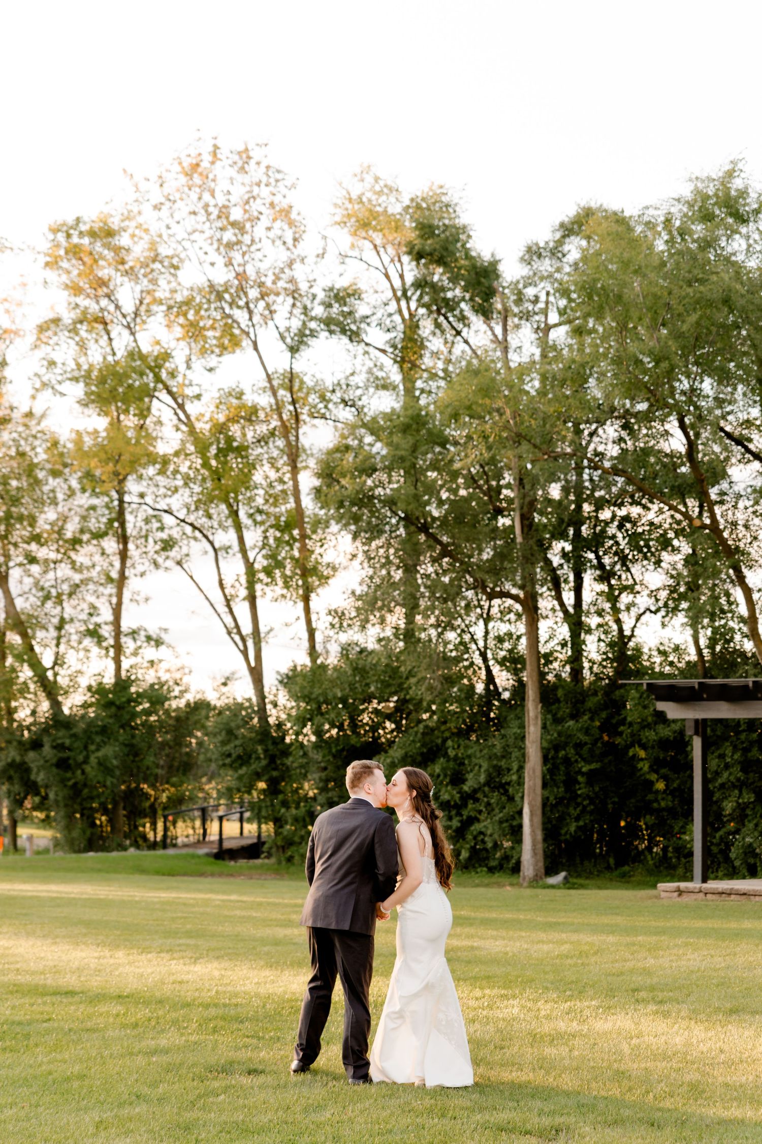 Bride and groom share a romantic kiss on a lush green lawn surrounded by tall trees during golden hour.