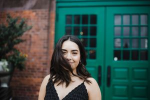A person in a black dress poses against a brick wall with bright teal windows in an outdoor photoshoot setting.