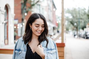 Woman in denim jacket smiles and laughs while walking down urban sidewalk on sunny day.