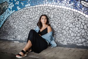 A person in a black dress and denim jacket sits against a mosaic wall with a blue and white pattern.