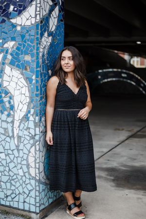 Stylish poses near a blue mosaic column in a parking structure wearing a flowing black dress.