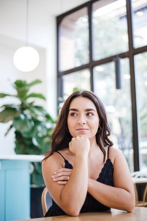 A woman in a black tank top leans on a table near large windows and indoor plants in a bright modern space.