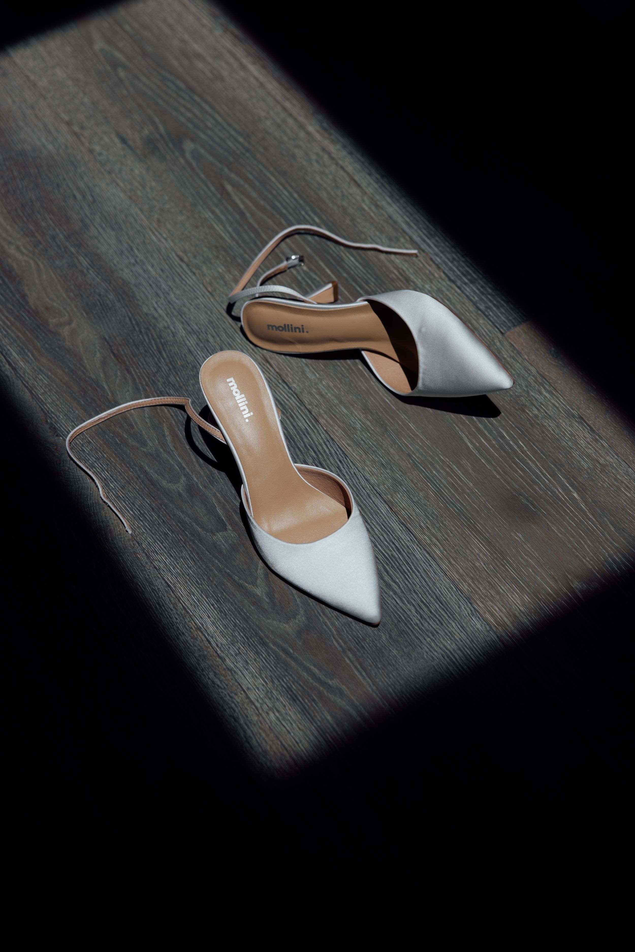 White pointed-toe bridal shoes on dark wooden flooring with dramatic lighting.