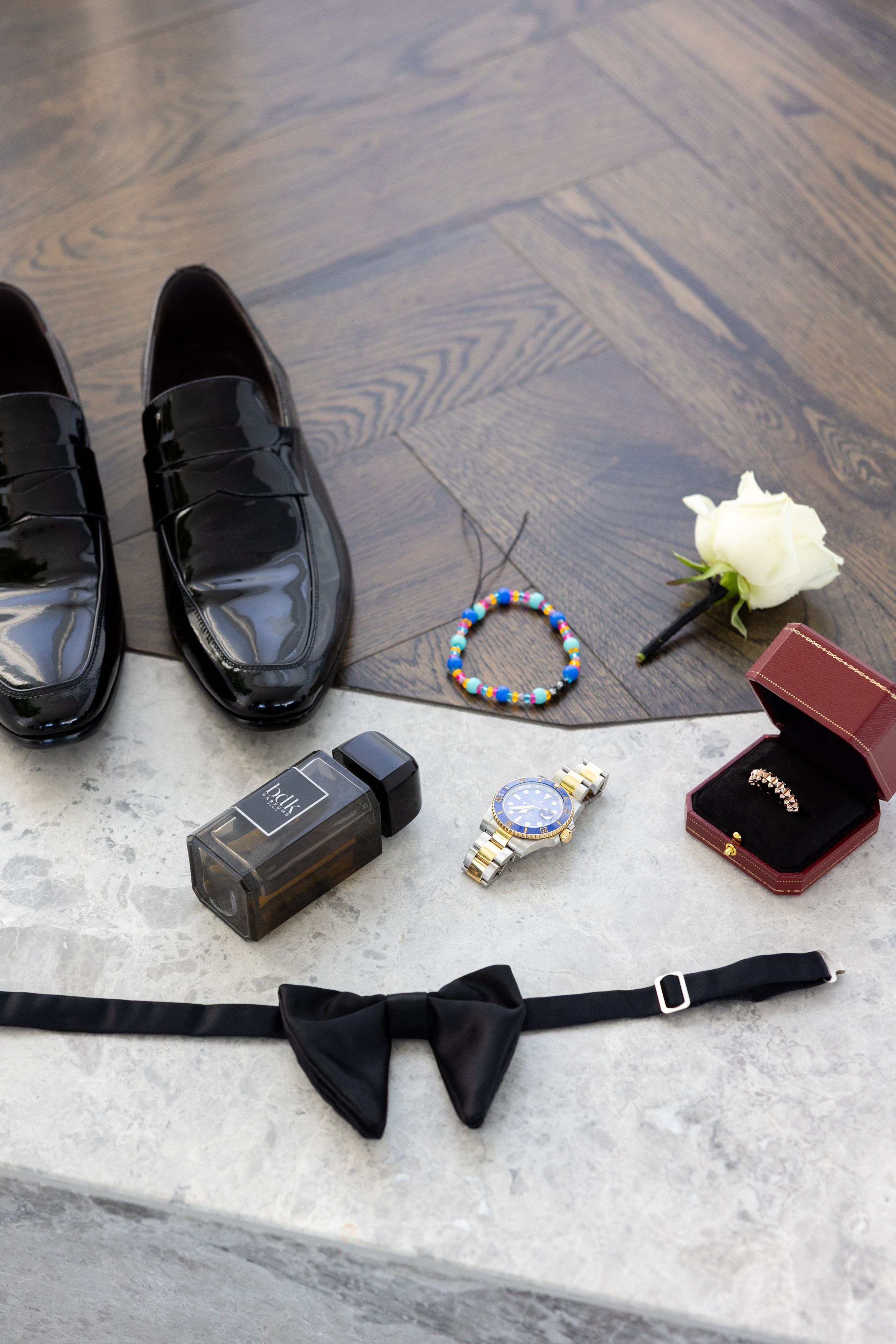 Wedding accessories including black dress shoes, bow tie, watch, ring box and white rose on wooden floor.